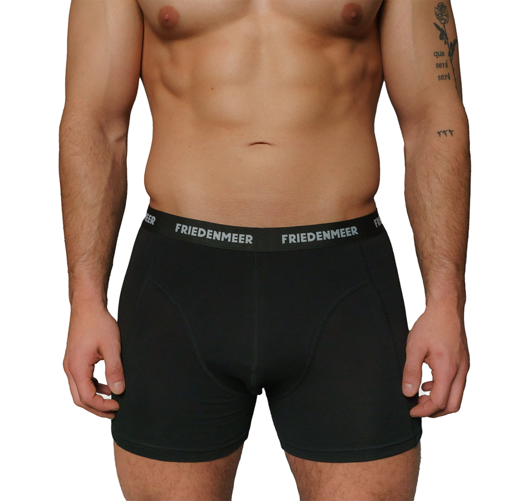 The image depicts the upper body of an athletic man showcasing and wearing the black boxer shorts by FRIEDENMEER, featuring a white 'FRIEDENMEER' text design on a black waistband, showcased and presented.