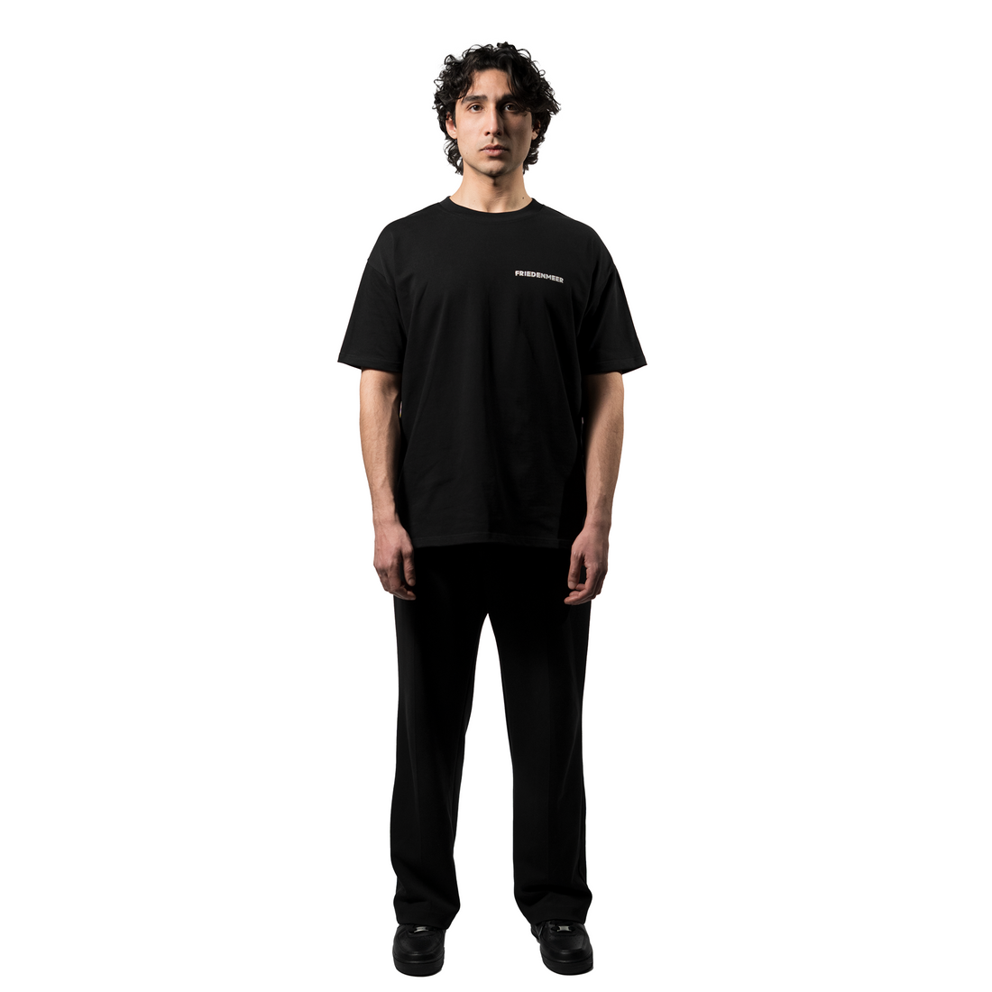 A model showcases an elegant oversized black T-shirt with the white brand name FRIEDENMEER across the chest, paired with matching black trousers and shoes, highlighting the brand's sleek elegance luxury.