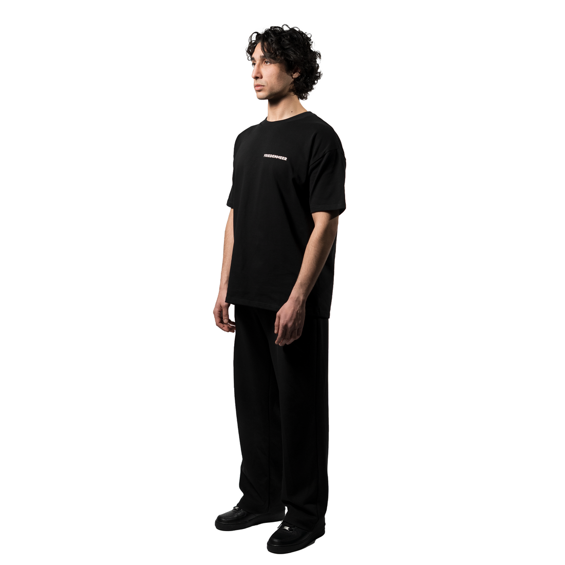 A model showcases an elegant oversized black T-shirt with the white brand name FRIEDENMEER across the chest, paired with matching black trousers and shoes, highlighting the brand's sleek elegance luxury.